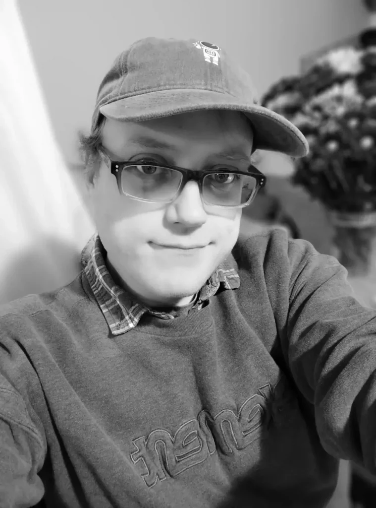 Justin-Stanelle-BW Black-and-white selfie of Justin Stanelle wearing black-framed glasses and a cap with a minimalist robot logo, smiling slightly with a casual plaid shirt and sweatshirt, with blurred flowers in the background.